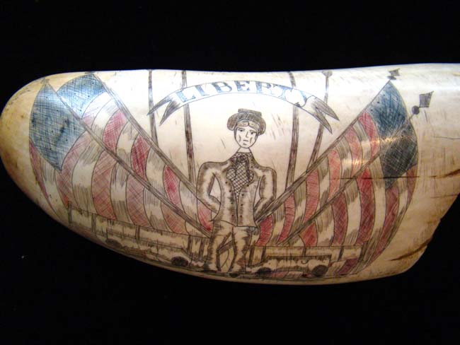 An attractive 'Liberty' themed 'sag harbor' style scrimshawed whale's tooth depicting a soldier flanked by a pair of cannons with four American flags in the background